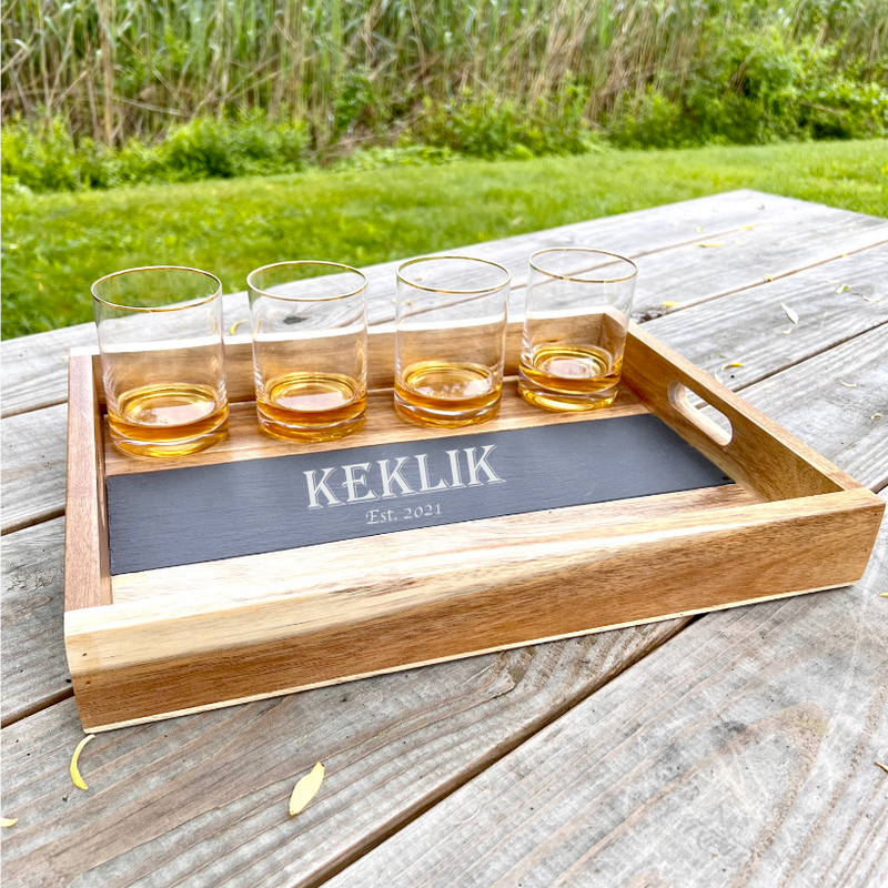 Wooden serving tray with four whiskey glasses filled with amber liquid, featuring a removable slate insert engraved with sample name "KEKLIK" and "Est. 2021