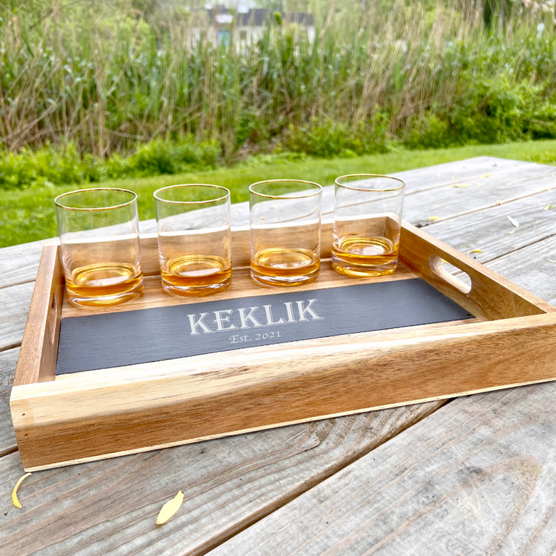 Wooden tray with removable slate insert engraved with sample name "KEKLIK" and "Est. 2021", holding four gold-rimmed whiskey glasses filled with amber liquid, displayed on weathered deck with garden background