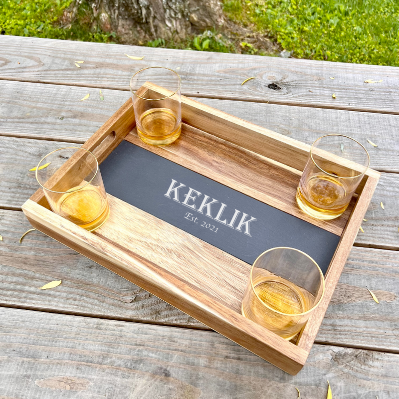 Wooden acacia tray with removable slate insert engraved with sample name "KEKLIK" and "Est. 2021," styled with four gold-rimmed whiskey glasses containing amber liquid.