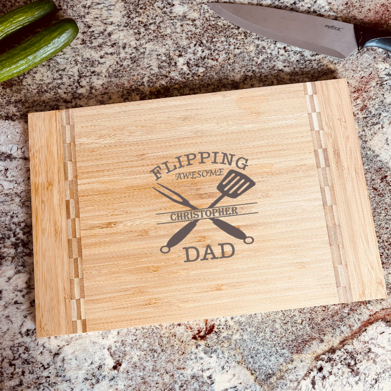 Bamboo cutting board engraved with sample name "Christopher" integrated into "Flipping Awesome Dad" design with crossed spatula and knife, displayed on granite countertop