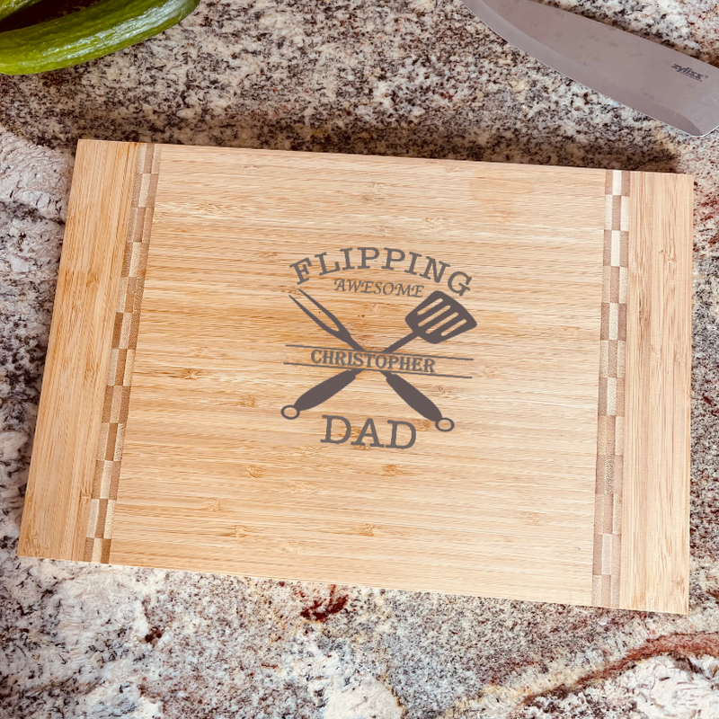 Bamboo cutting board engraved with sample name "Christopher" integrated into "Flipping Awesome Dad" design with crossed spatula and knife icons, displayed on granite countertop