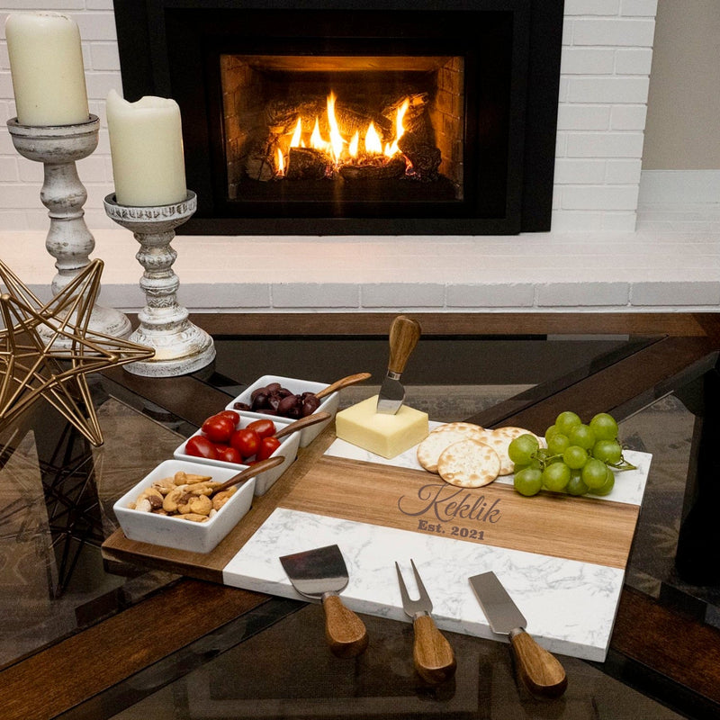 Wooden and marble charcuterie board engraved with sample name "Keklik" and date "Est. 2021" styled with cheese, crackers, grapes, and cured meats near a fireplace