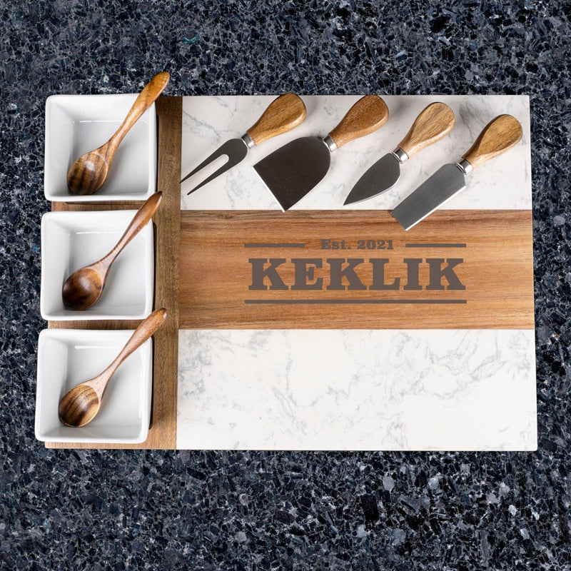 Engraved charcuterie board with sample name "KEKLIK" and establishment date "Est. 2021" on wood strip, displayed with five cheese knives and three wooden serving spoons in white ceramic bowls.