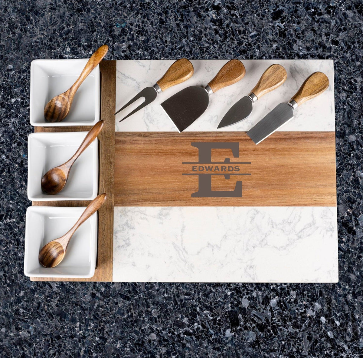 Overhead view of acacia wood and marble charcuterie board engraved with sample initial "E" and name "EDWARDS," displayed with cheese knives and wooden serving spoons in white ceramic dishes.