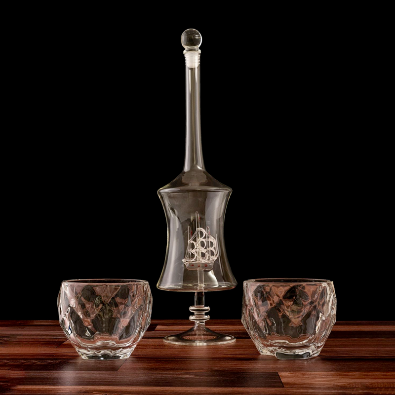Tall glass decanter with cork stopper and engraved sailship design, flanked by two textured whiskey glasses on wooden surface