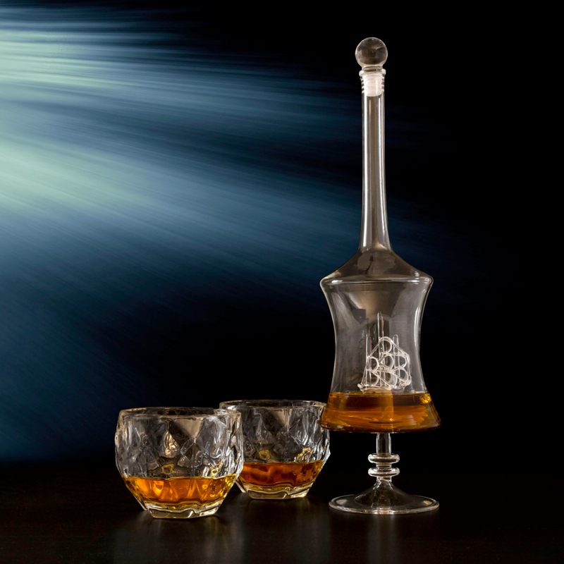Tall glass decanter with handcrafted ship design inside, filled with amber whiskey, displayed with two matching weighted rocks glasses containing whiskey and ice.