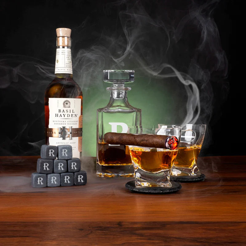 Whiskey Crate Gift Set displayed with bourbon bottle, green glass decanter, two glasses filled with amber liquid, cigar, personalized whiskey stones engraved with sample letter "R", and slate coasters on wooden table.
