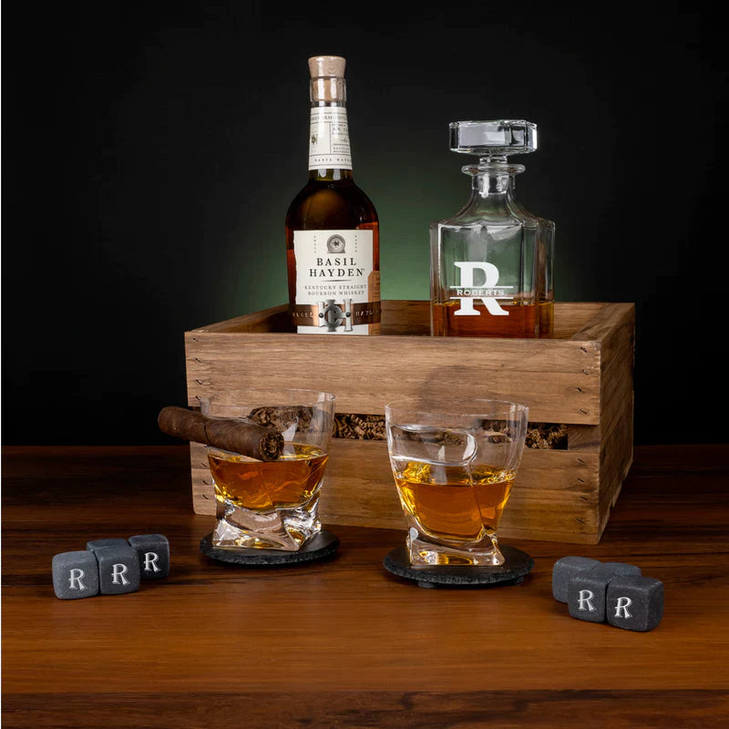 Wooden crate whiskey set displayed with decanter engraved with sample initial "R", two rocks glasses with cigar notches filled with whiskey, slate coasters and whiskey stones each engraved with sample letter "R", and a cigar on dark wood surface.