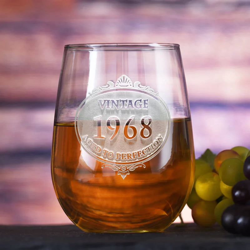Stemless wine glass engraved with "Vintage 1968 Aged to Perfection" in decorative circular medallion, filled with amber liquid, grapes beside on sunset background
