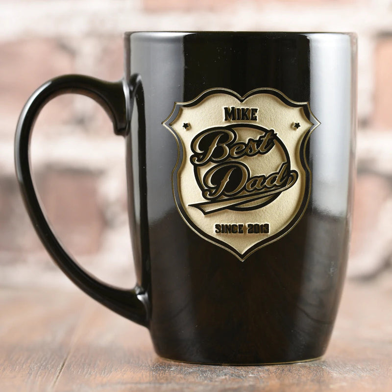 Black stoneware mug with engraved gold shield-shaped badge featuring sample name "MIKE," "Best Dad," and "SINCE 2013" in raised 3D relief lettering.