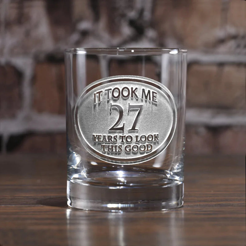Clear whiskey glass engraved with circular medal design reading "It Took Me 27 Years to Look This Good" on wooden surface against brick background