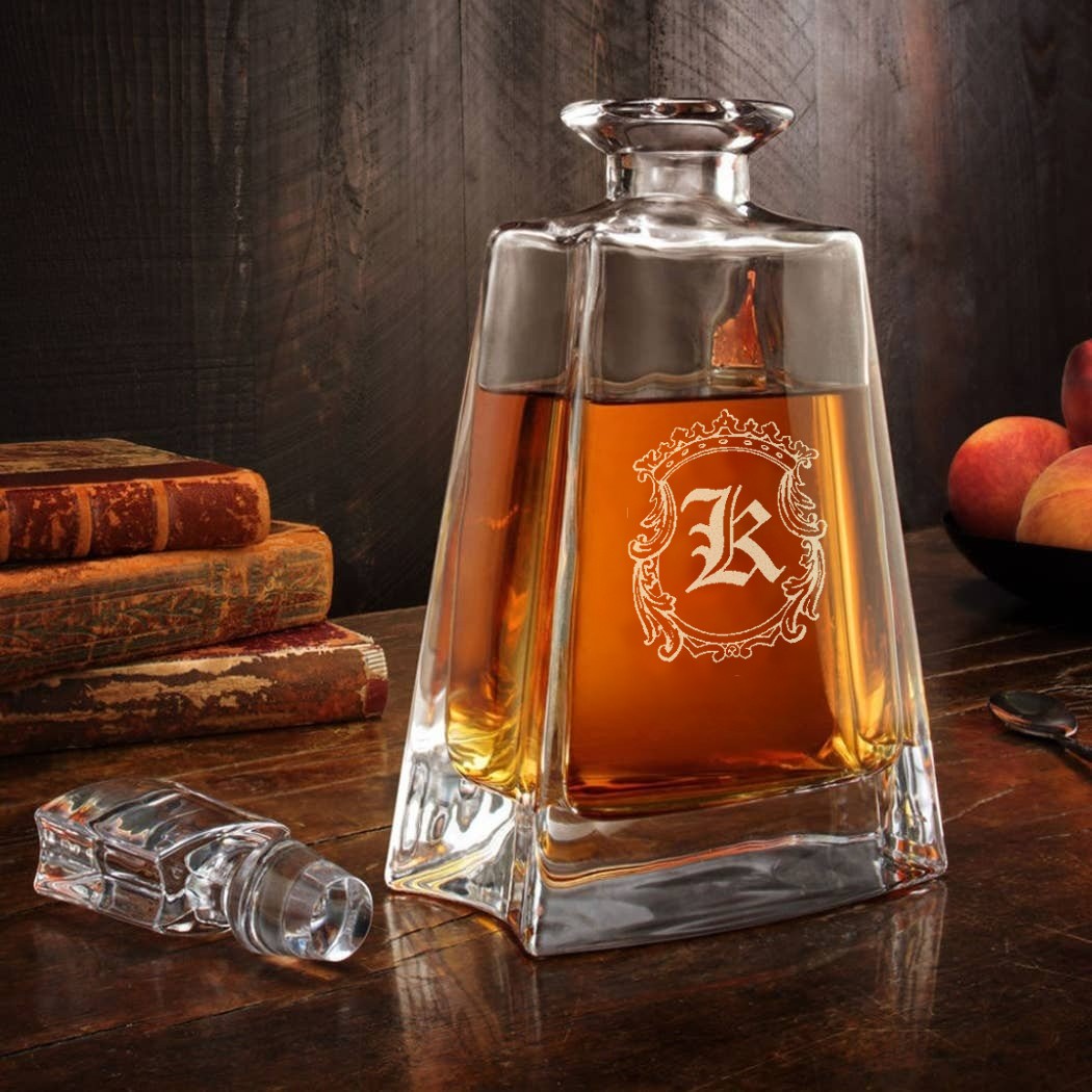 Crystal decanter filled with amber whiskey, engraved with ornate frame around sample monogram K, displayed with books and matching stopper on dark wood surface