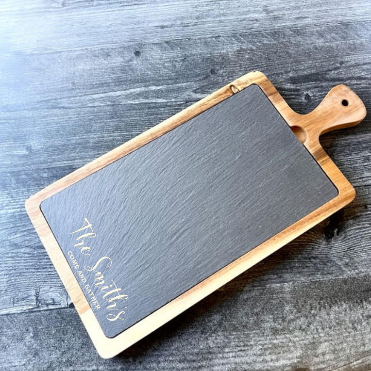 Slate and acacia wood serving board engraved with sample name "Smith" and date on the dark inlay surface, displayed on weathered wood background