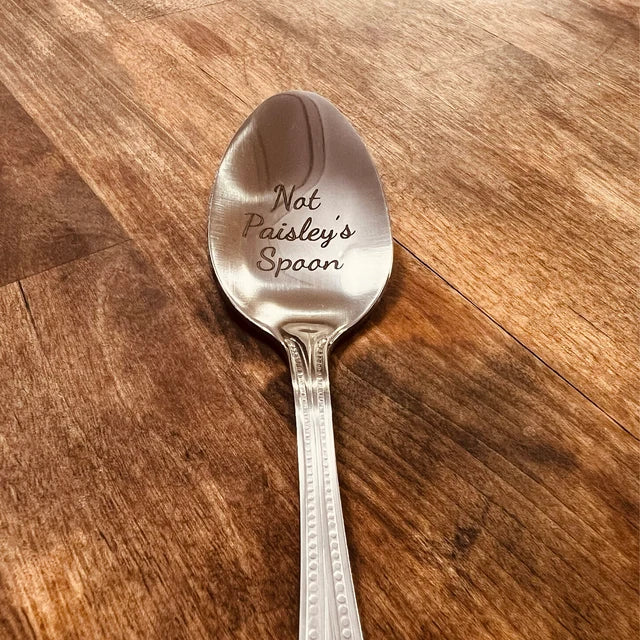 Stainless steel spoon engraved with sample text "Nat Paisley's Spoon" on the bowl, displayed on a wooden surface