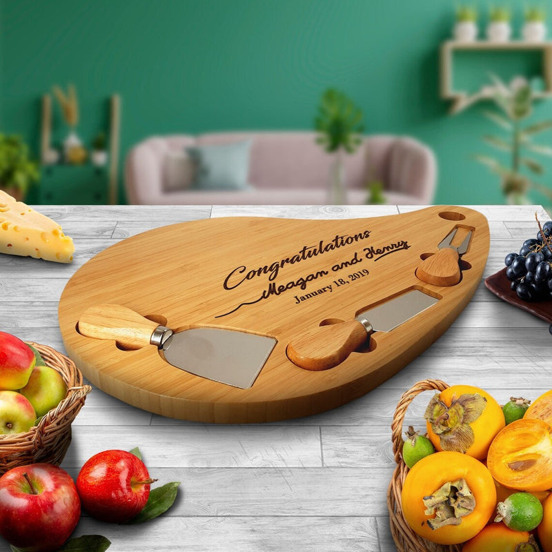 Bamboo teardrop cheese board engraved with sample text "Congratulations Megan and Henry" and date "January 18, 2019", featuring two metal-inset cheese knives and hanging hole, displayed on white wood table surrounded by fresh fruit and cheese.
