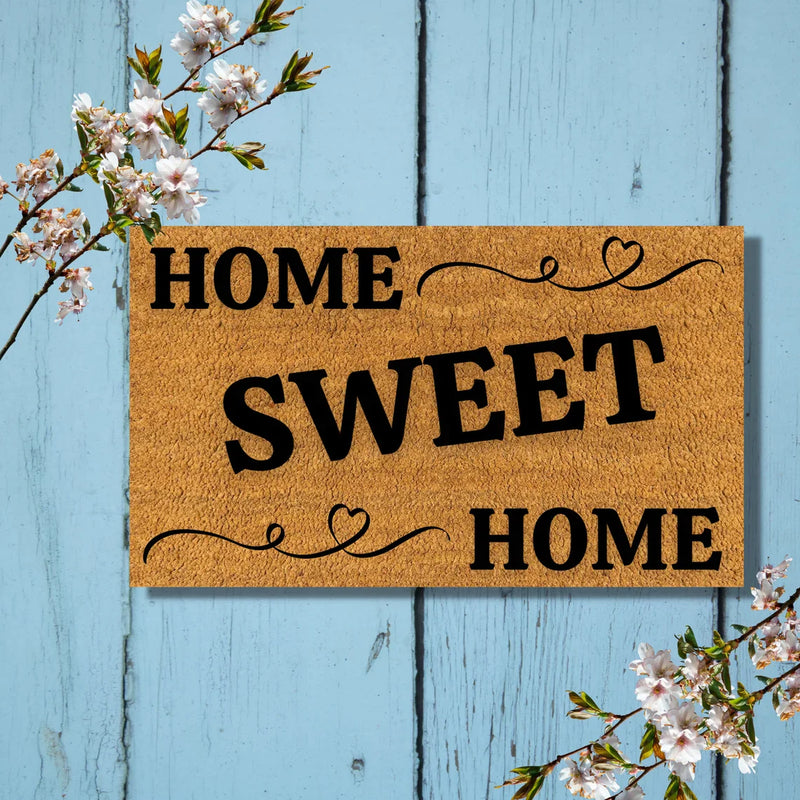 Tan coir doormat printed with "HOME SWEET HOME" in black text with decorative heart flourishes, displayed on light blue wooden planks with white flowering branches.