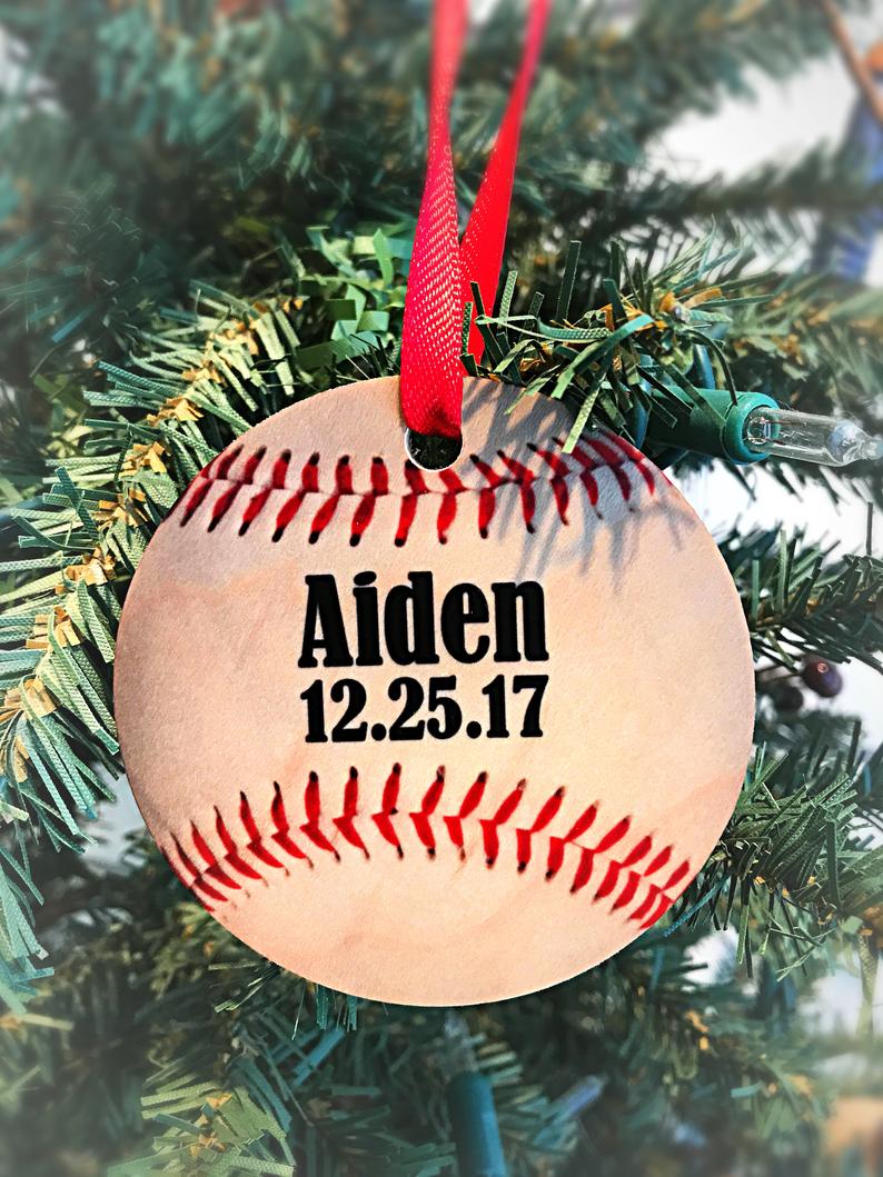 Wooden baseball ornament with red stitching hanging from red ribbon on evergreen branch, personalized with sample name "Aiden" and date "12.25.17