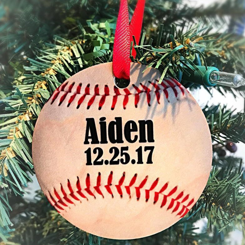 Wooden baseball ornament with red silk thread stitching, personalized with sample name "Aiden" and date "12.25.17", hanging from red ribbon on evergreen branch