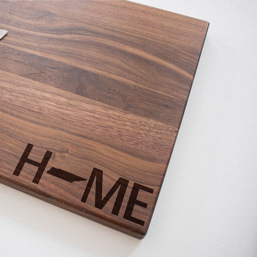 Walnut hardwood cutting board with laser-engraved "HOME" text in the lower corner, angled view showing wood grain and finish