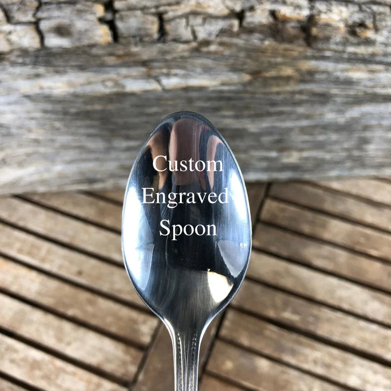 Polished stainless steel spoon with bowl facing up, displayed against weathered wood background, showing reflective surface ready for custom engraving