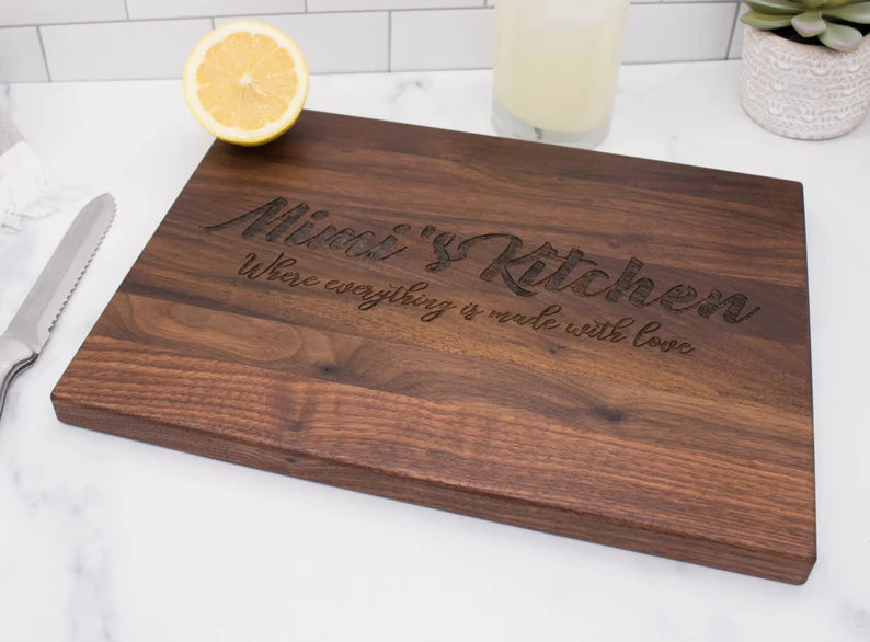 Dark walnut cutting board engraved with sample text "Alfred's Kitchen" and "Where everything is made with love" displayed on marble countertop with lemon and beverage