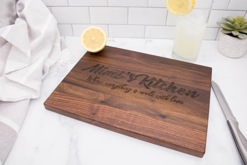 Walnut cutting board engraved with sample text "Blessed Kitchen" and "Where everything is made with love" displayed on white marble with lemon slices and a glass of lemonade nearby