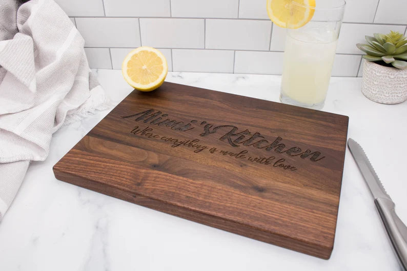 Dark walnut cutting board engraved with sample text "Happy Kitchen" and script lettering, displayed on marble countertop with lemon water and kitchen linens
