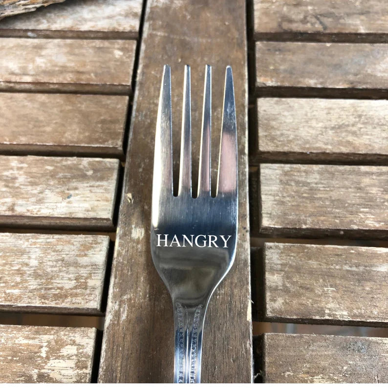 Stainless steel fork engraved with sample text "HANGRY" on the handle, displayed on weathered wood surface