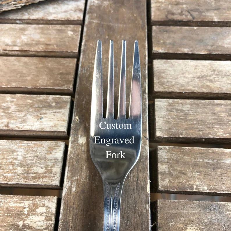 Stainless steel fork engraved with sample text "Custom Engraved Fork" on the handle, displayed on weathered wooden surface
