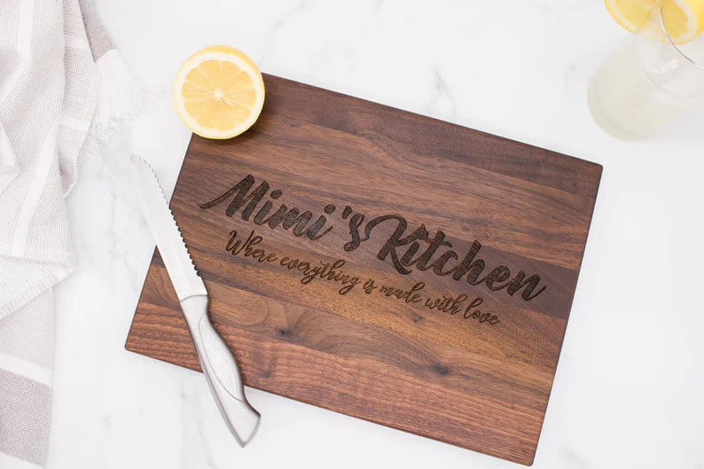 Dark walnut cutting board engraved with sample text "Nuni's Kitchen" and "Where everything is made with Love," styled with lemon slices and a glass of water
