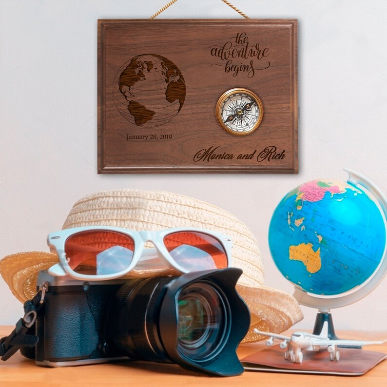 Wooden plaque with engraved globe, "the adventure begins" text, sample names "Monica and Rich," date "January 28, 2019," and attached brass compass, displayed with travel-themed props including camera, sunglasses, and globe.