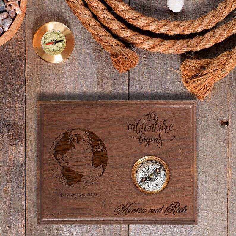 Wooden plaque engraved with sample names "Monica and Rich," engagement date "January 28, 2019," globe design, and "the adventure begins" text, decorated with brass compass inset