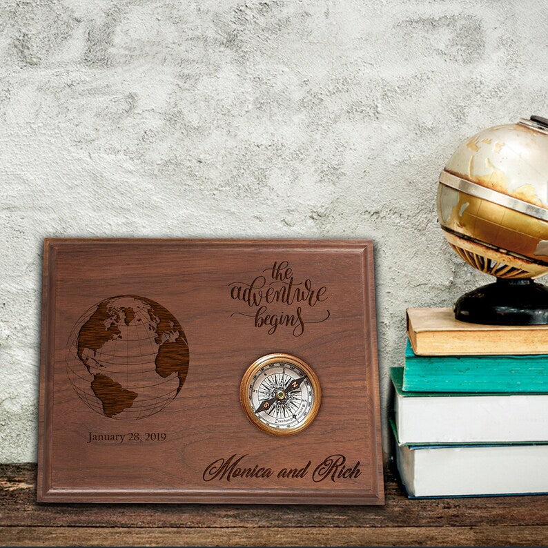 Walnut wood plaque engraved with globe design, "the adventure begins" text, sample names "Monica and Rich," date "January 28, 2019," and embedded brass compass