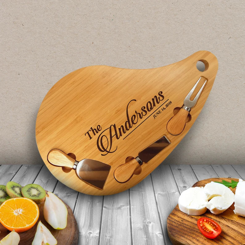 Teardrop-shaped bamboo charcuterie board engraved with sample name "The Andersons" and date "June 14, 2018", displayed with three attached cheese knives and fresh fruit on wooden surface.