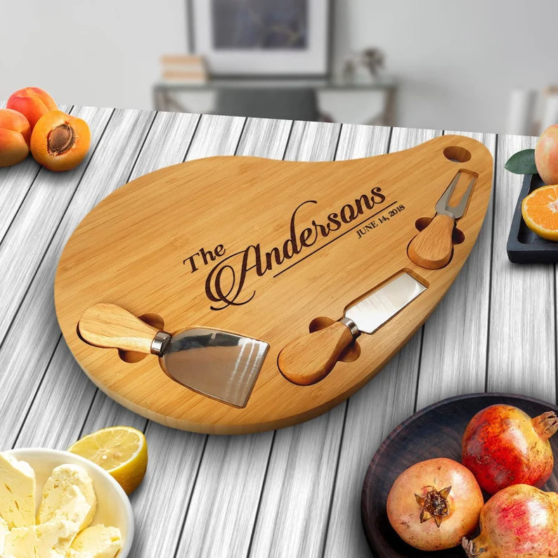 Teardrop-shaped bamboo charcuterie board engraved with sample name "The Andersons" and date "June 24, 2018", featuring three attached cheese knives with wooden handles, displayed on white wood table with fresh citrus and pomegranates