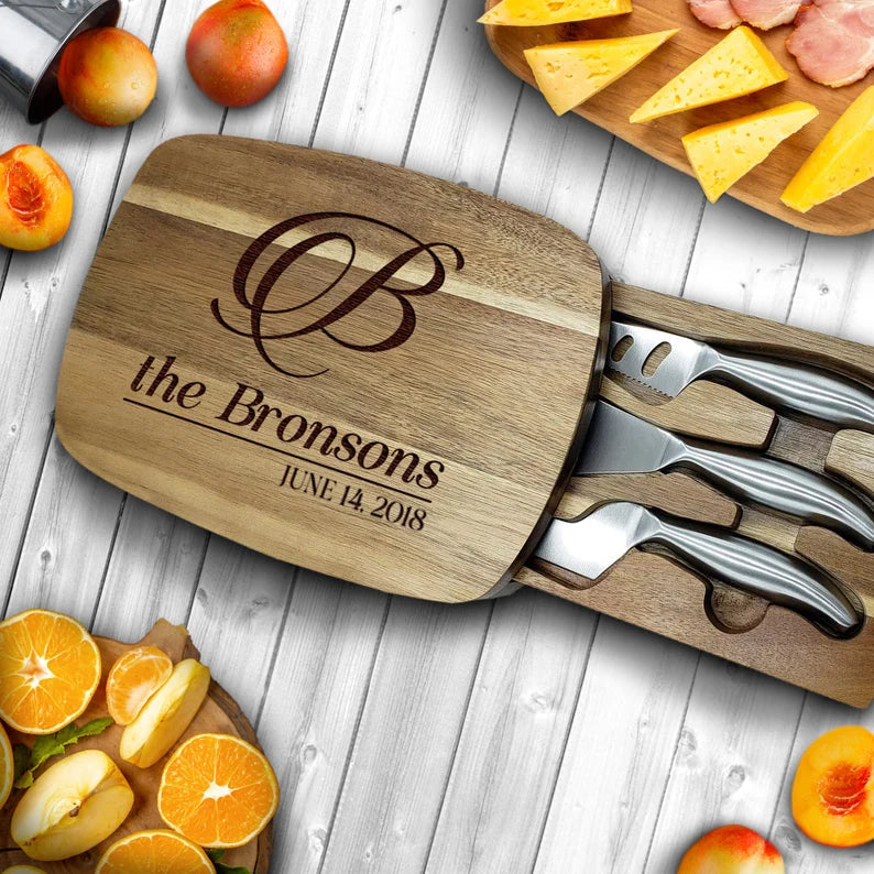 Acacia wood charcuterie board engraved with decorative monogram, "the Bronsons," and sample date "JUNE 14 2018," displayed with knife set and fresh fruit on white wood surface.
