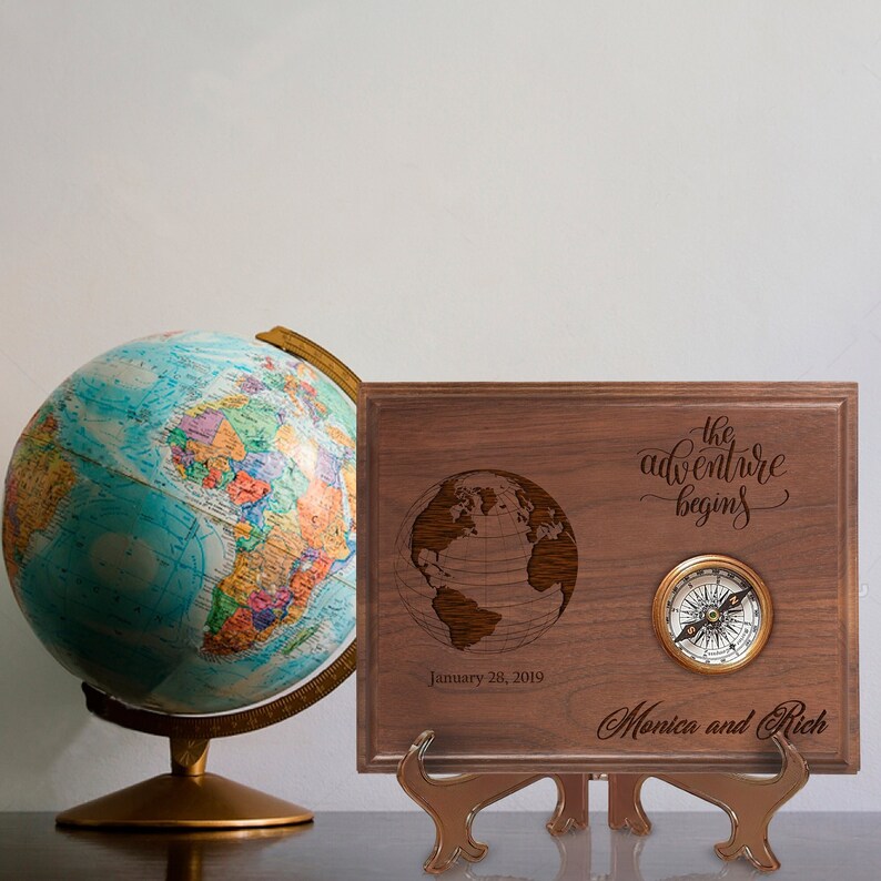 Walnut plaque engraved with "the adventure begins" text, world globe illustration, sample date "January 28, 2019", and sample names "Monica and Rich", displayed with decorative brass compass and standing easel next to colorful desktop globe.