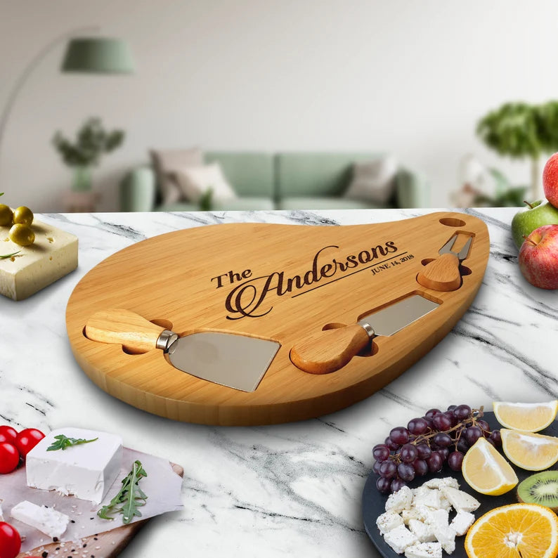 Bamboo teardrop charcuterie board with sample engraving reading "The Andersons" and a date, styled on marble countertop with fresh produce and cheese