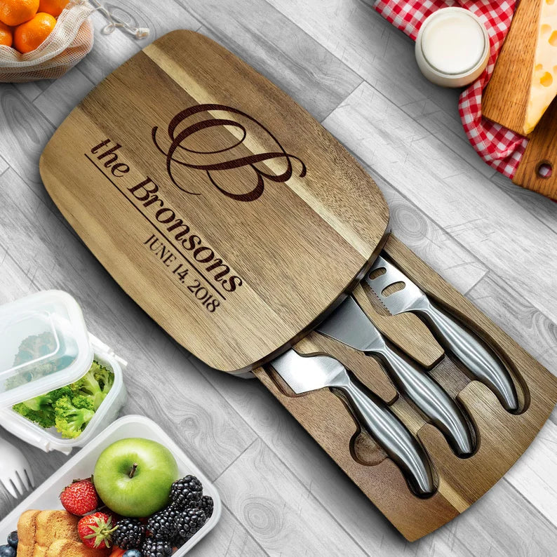 Wooden charcuterie board engraved with sample name "the Bronsons" and date "JUNE 4, 2018," featuring four stainless steel serving utensils with black handles, displayed on gray wood surface with fresh fruit and cheese nearby.