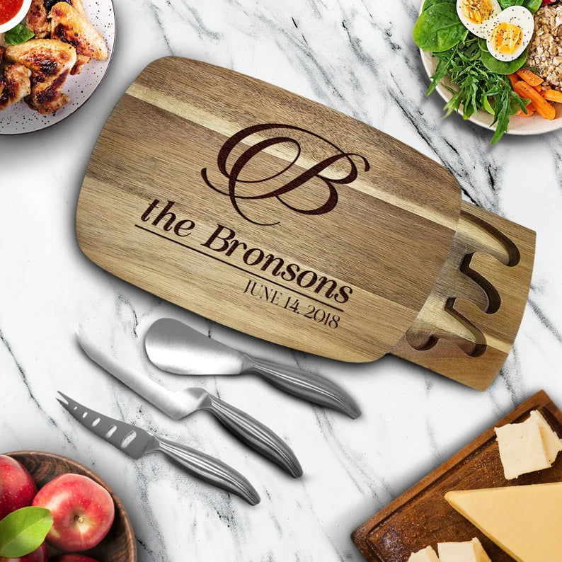 Acacia wood charcuterie board engraved with decorative monogram, "the Bronsons," and sample date "JUNE 14 2018," displayed with cheese knives and fresh food styling on marble surface