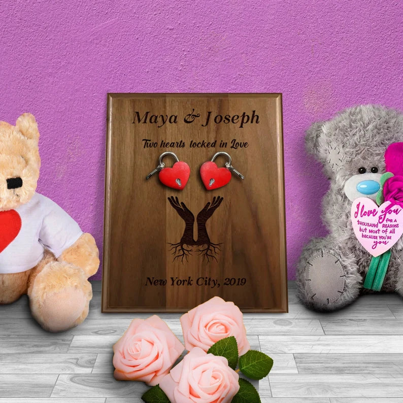 Walnut wood plaque engraved with sample names "Maya & Joseph" and text "Two hearts locked in Love," decorated with red heart-shaped padlocks and displayed with pink roses and plush teddy bears.