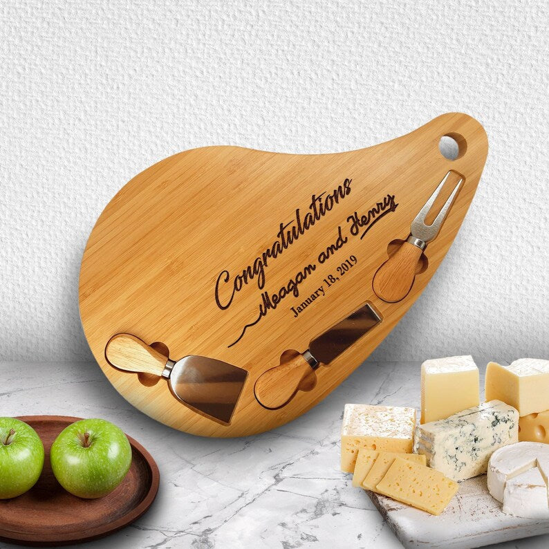 Bamboo teardrop cheese board engraved with sample text "Congratulations Megan and Henry January 18, 2019," displayed with wooden serving knives and assorted cheeses and apples
