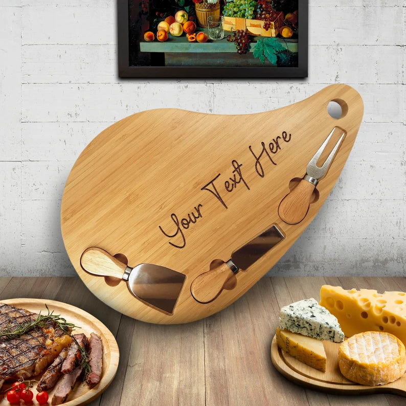 Bamboo teardrop charcuterie board with three attached cheese knives, engraved with sample text "Your Veg Here", displayed with sliced meats and assorted cheeses
