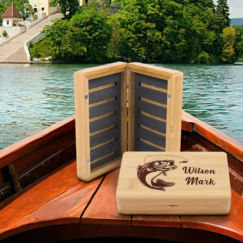 Bamboo fly fishing tackle box with engraved sample name "Wilson Mark" and fish illustration, displayed open on a wooden boat with fly case organizer in background and lake scenery