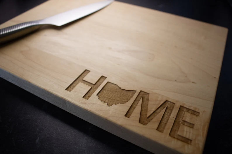 Wooden cutting board laser-engraved with "HOME" text and sample state outline of Ohio in the center, metal knife visible at top edge