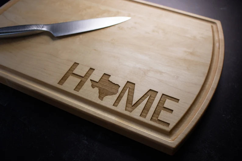 Hardwood cutting board with laser-engraved "HOME" text and Texas state outline, shown with a chef's knife resting on the surface