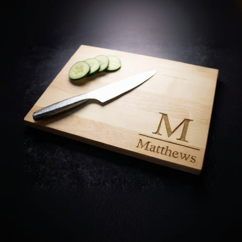 Light wood cutting board engraved with sample monogram "M" and name "Matthews," displayed with sliced cucumber and a kitchen knife