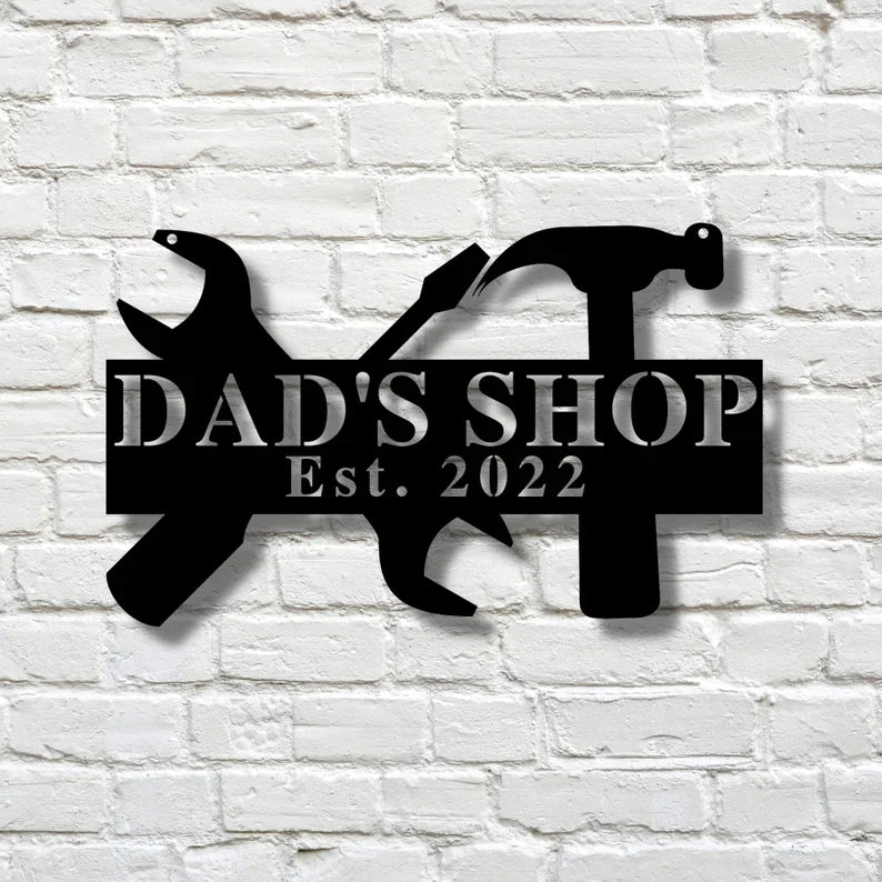 Black laser-cut steel workshop sign with wrench and hammer silhouettes, personalized with sample text "DAD'S SHOP Est. 2022" mounted on natural wood background.