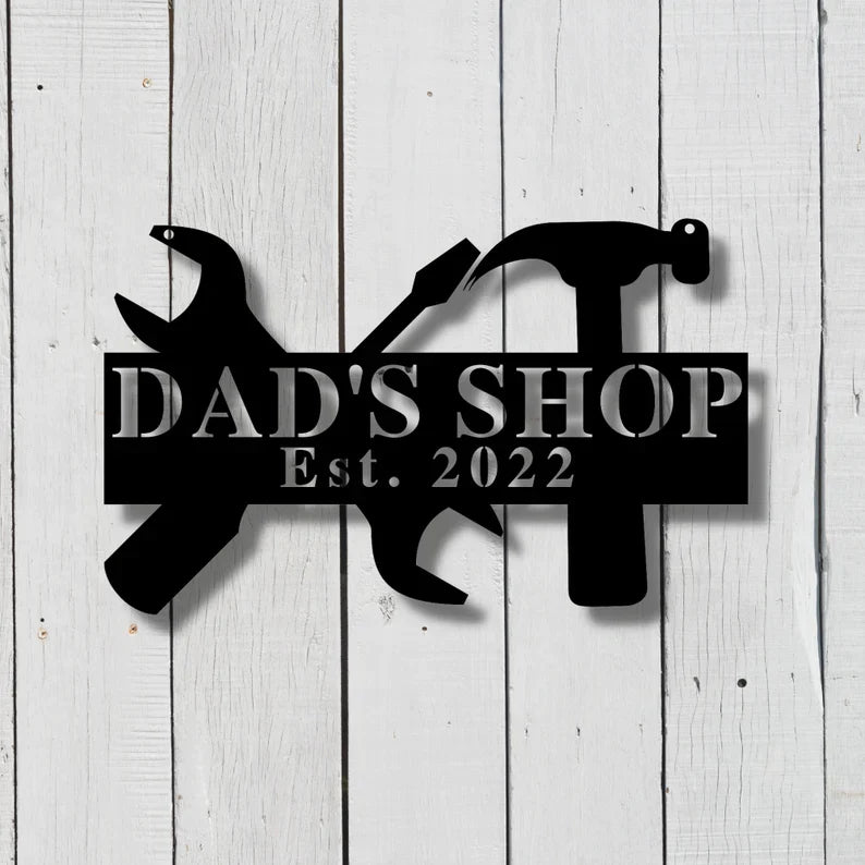 Black laser-cut metal workshop sign with sample personalization reading "DAD'S SHOP Est. 2022" and tool silhouettes, mounted on white shiplap wall