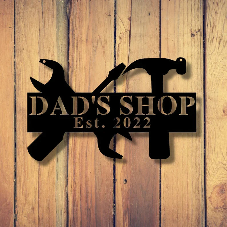 Black laser-cut steel workshop sign with wrench and hammer silhouettes, personalized with sample text "DAD'S SHOP Est. 2022" mounted on natural wood background.